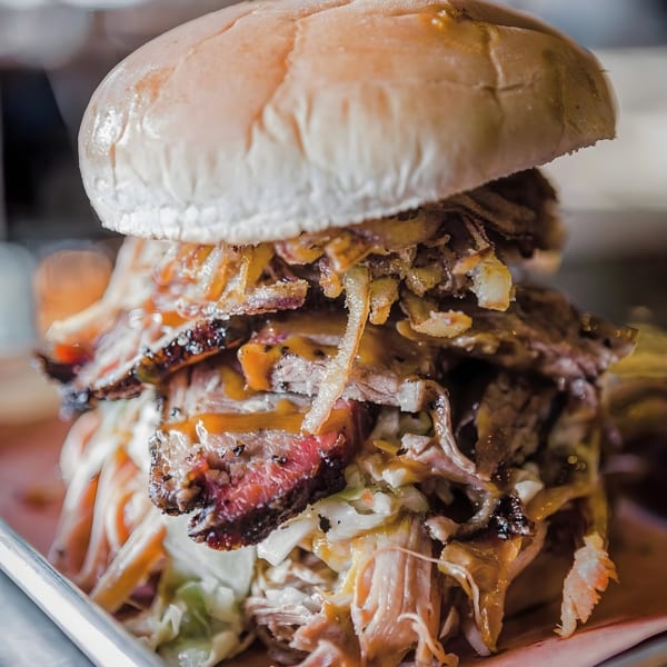 Close-up of a Bessinger's BBQ sandwich overflowing with pulled pork, brisket, coleslaw, and fried onions.