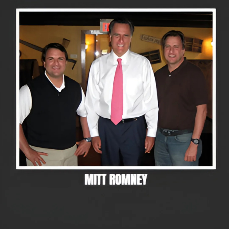 Mitt Romney in a white shirt and pink tie, flanked by two men, inside Bessinger's BBQ.