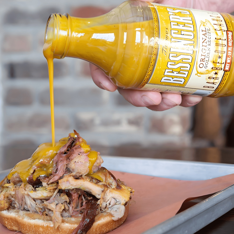 A hand pours Bessinger's golden barbecue sauce from a bottle onto a pulled pork sandwich.