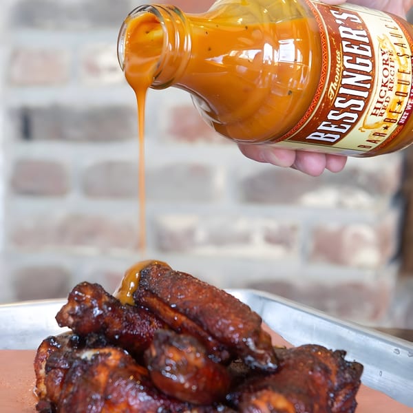 A hand pours Bessinger's BBQ sauce from a bottle onto a tray of dark, crispy chicken wings.