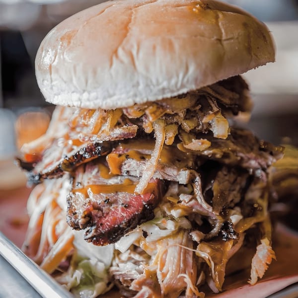 A towering Bessinger's BBQ sandwich, overflowing with smoked meats, coleslaw, and sauce on a bun.