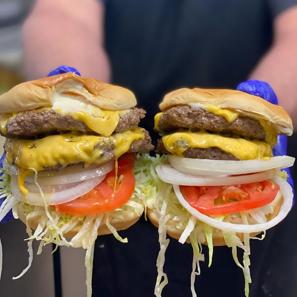 Two double cheeseburgers with melted cheese, lettuce, tomato, and onion, held by gloved hands.