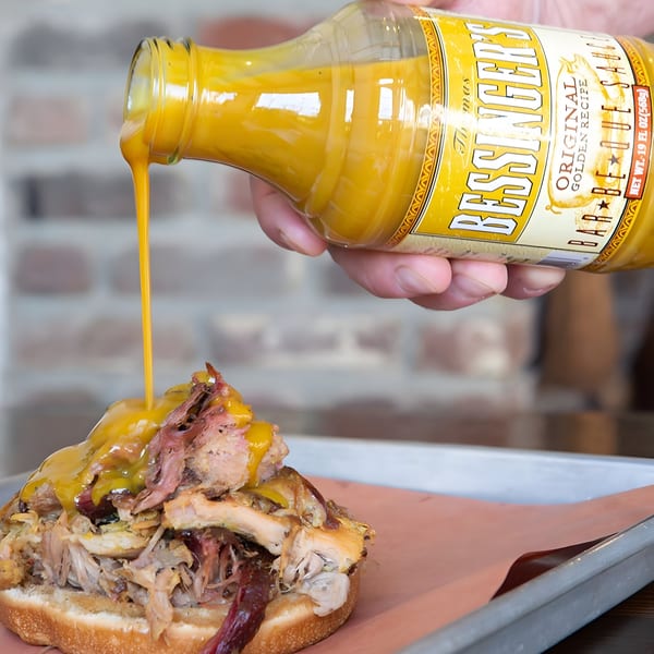 A hand pours Bessinger's golden barbecue sauce from a bottle onto a pulled pork sandwich.