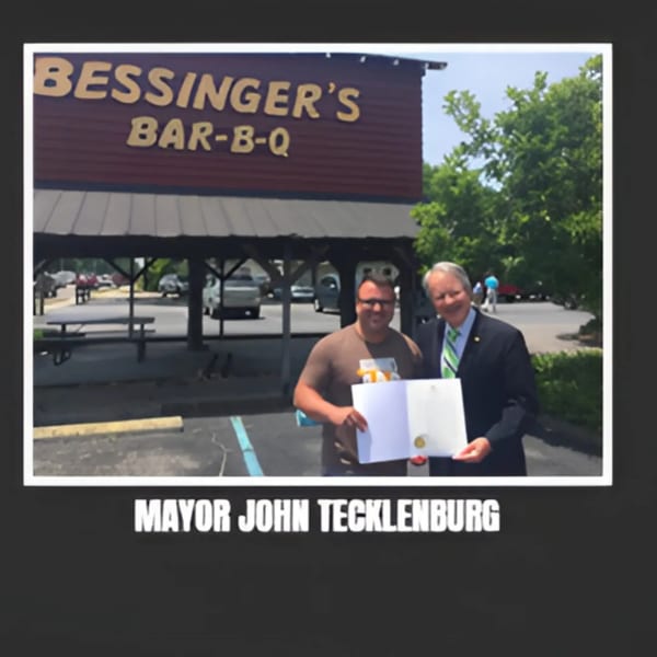 Two men, one in a suit, holding a document in front of Bessinger's BBQ restaurant.