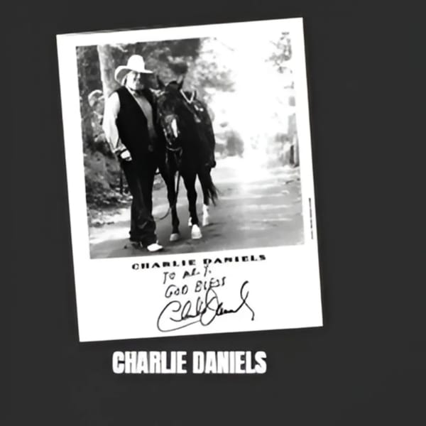 Black and white photo of Charlie Daniels in a cowboy hat, walking a horse, with his autograph.