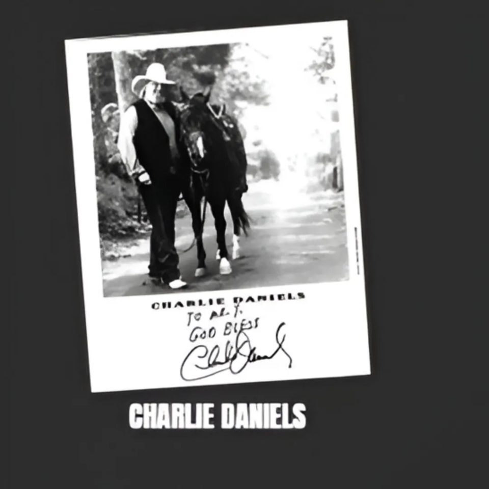 Black and white photo of Charlie Daniels in a cowboy hat, walking a horse, with his autograph.
